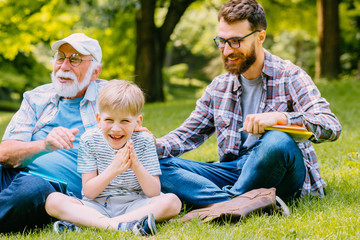 happy family of three generation - father, grandfather and blond son sitting on grass at park with books learn to read while getting ready for school.They are laugh,fun,spending good time together
