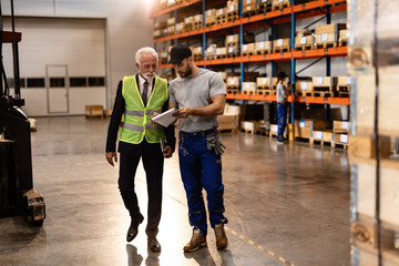 Senior manager and young worker going through paperwork while walking through a warehouse.