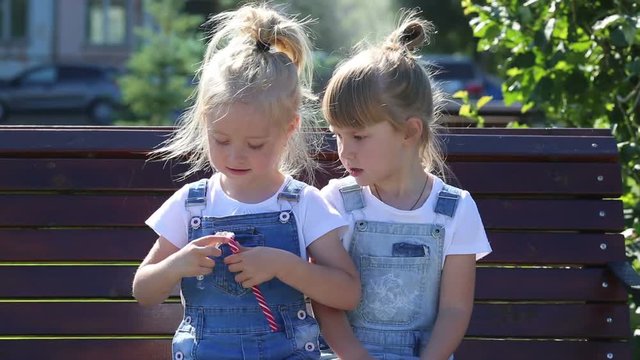 The girl shares candy with her little friend. Two girls of 5 years old are sitting on a bench in the park and they are breaking up a big candy. One girl treats another.