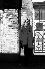 A beautiful blonde girl wearing a red coat, sunglasses and gloves, stands by a brick wall lit by the bright sun. Fashion, commercial, advertising design.  Black and white art photo. Copy space