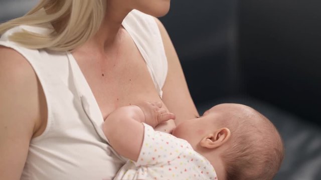 Young blonde breastfeeding mother is smiling while her baby is eating milk. Closeup. 
