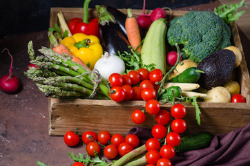 Box with farm vegetables on a dark background. Place for text. Cherry tomatoes, asparagus, broccoli, peppers, eggplants, zucchini, carrots and onions.