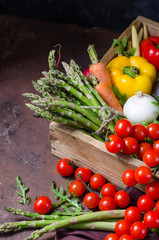 Box with farm vegetables on a dark background. Place for text. Cherry tomatoes, asparagus, broccoli, peppers, eggplants, zucchini, carrots and onions.