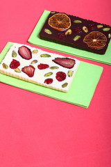 Homemade white and black chocolate. Decorated with slices of dried orange, strawberries, cherries and pistachios. It lies on a light green napkin on a coral background.