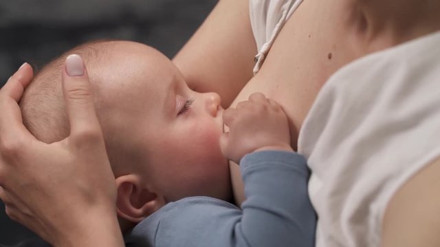 Close-up shot of mother breastfeeding her sleepy baby. Baby is touching the breast. Mother pats on the head. 