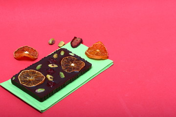 Homemade black chocolate. Decorated with slices of dried orange, strawberries, cherries and pistachios. It lies on a light green napkin on a coral background.