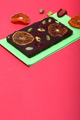 Homemade black chocolate. Decorated with slices of dried orange, strawberries, cherries and pistachios. It lies on a light green napkin on a coral background.