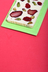 Homemade white chocolate. Decorated with slices of dried strawberries, cherries and pistachios. It lies on a light green napkin on a coral background.