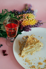 Photo of a beautifull delicious slice of homemade layered cake on a porcelain white plate with a glass of fresh lemonade and color flowers on pink background indoor rustic style