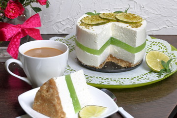 Lime cheesecake without baking. The piece is cut off. Lies on a plate. Near a cup of coffee with milk. In a glass vase is a bouquet of roses.