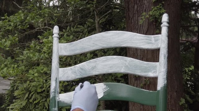 Slow motion view of a hand wearing latex glove is painting the back of a green wooden chair white with a brush. This is being done outside with a tree and leaves in the background