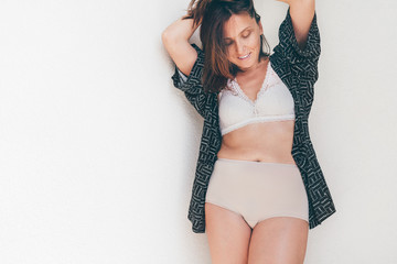 Middle-aged Caucasian woman in light tones bra and panties underwear posing near light background...