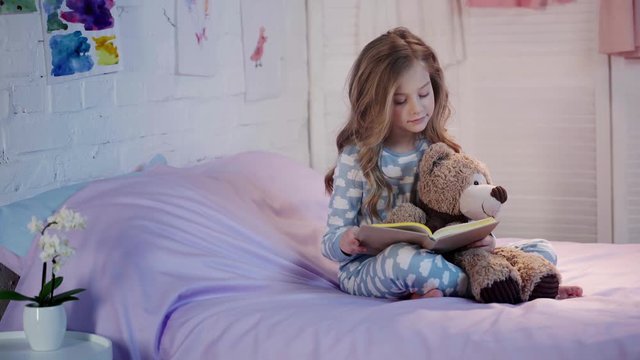 cute preteen child in pajamas sitting on bed with teddy bear, reading book, turning page and smiling in bedroom