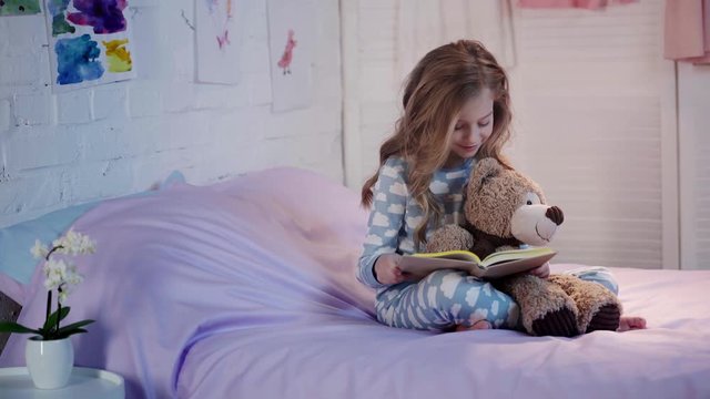 cute preteen child in pajamas sitting on bed with teddy bear, reading book, smiling and expressing amazement