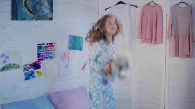 happy kid in pajamas holding teddy bear and jumping on bed in bedroom