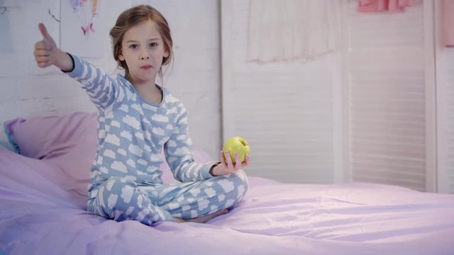 kid in pajamas sitting on bed, eating green apple, looking at camera and showing thumb up with smile