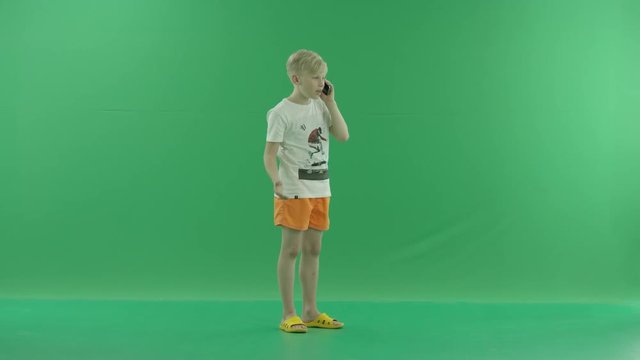 Angry young blonde boy in casual clothes is talking on his mobile phone. He stands sideways against a greenscreen