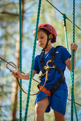 Child in forest adventure park. Kid in red helmet and blue t- shirt climbs on high rope trail. Agility skills and climbing outdoor amusement center for children.