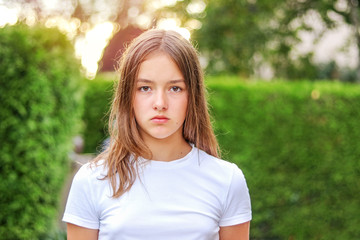 Close-up portrait of serious angry teenager girl outdoors in park with green hedge background. No...