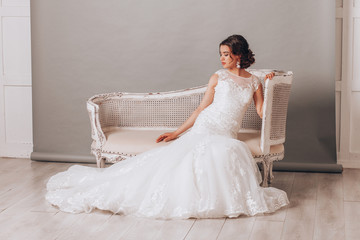 Young beautiful bride in a luxurious wedding dress sitting in a wicker chair in the studio on a gray background