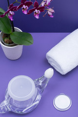 Obraz na płótnie Canvas Neti Pot with Soft Comfort Tip, pile of Saline, Purple Orchid Flowers and rolled up White Towels on purple background. Sinus wash. Nasal irrigation.