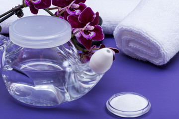 Obraz na płótnie Canvas Spa purple background with Neti Pot, pile of Saline, rolled up White Towels, stacked Basalt Stones and Orchid Flower. Sinus wash. Nasal irrigation.