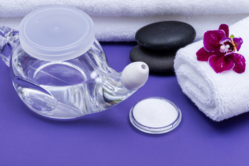 Fototapeta na wymiar Spa purple background with Neti Pot, pile of Saline, rolled up White Towels, stacked Basalt Stones and Orchid Flower. Sinus wash. Nasal irrigation.