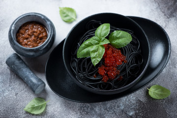 Bowl of squid-ink spaghetti with red caviar, sun dried tomato pesto and fresh green basil, elevated view on a grey stone background