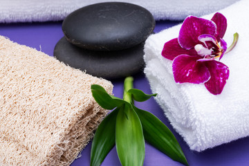 Fototapeta na wymiar Spa Wellness Concept. Natural Loofah Sponge, rolled up White Towels, stacked Basalt Stones, Bamboo and Orchid Flower on purple background.