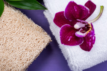 Obraz na płótnie Canvas Spa Wellness Concept. Natural Loofah Sponge, rolled up White Towels, stacked Basalt Stones, Bamboo and Orchid Flower on purple background.