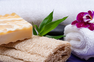 Obraz na płótnie Canvas Spa Wellness Concept. Natural Loofah Sponge, Almond Goat's milk Soap, White Towels, Basalt Stones, Bamboo and Orchid Flower on purple background.