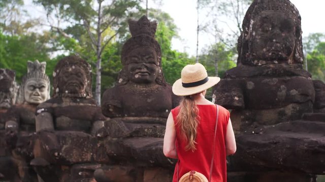 Female tourist is walking along the row of asuras and devas statues exploring their bricks. Bridge to Angkor Thom temple in Cambodia. Jungle is on the background