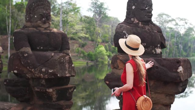Female tourist is walking along the row of asuras and devas statues exploring them. Bridge to Angkor Thom temple in Cambodia. River and jungle are on the background