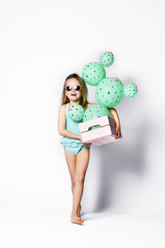 Young blonde girl in sunglasses and swimsuit holding a cactus ball in a pastel pink wooden box. The flowers are made of balloons with pink flowers and painted spines