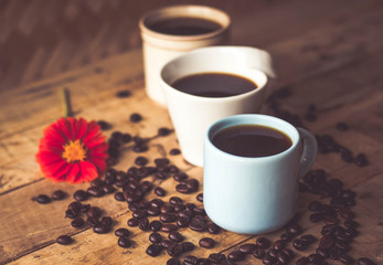 Black coffee In the cup sky blue color 1 and white 2 Have all 3, cups have roasted coffee beans are on table with, still have red flowers all placed on a wooden table.