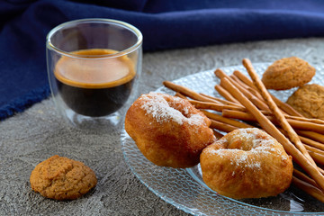 Two donuts on a plate, in the background of cup of espresso and blue kitchen towel.