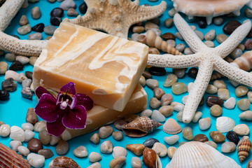 Fototapeta na wymiar Handmade ultra-moisturizing Almond Scented Goat's Milk Bar Soap decorated with Pebbles, Sea Stars, Sea Shells and Orchid Flower on blue background.