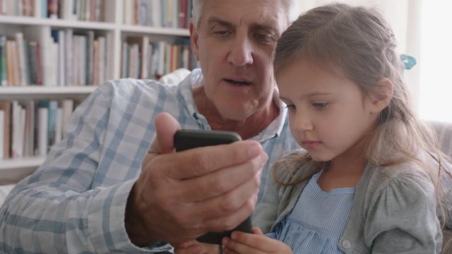 happy grandfather showing little girl how to use smartphone teaching curious granddaughter modern technology intelligent child learning mobile phone sitting with grandpa on sofa