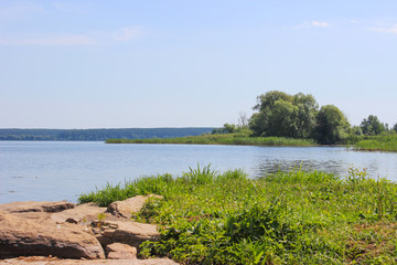 Shore of the reservoir with thick green grass. Near big stones of gray,brown color. On the opposite shore, thickets and trees. The blue surface of the water which reflects the high sky.