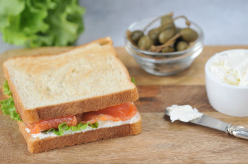 Salmon sandwich on a light wooden board. Cups with capers and soft cheese. Close-up.