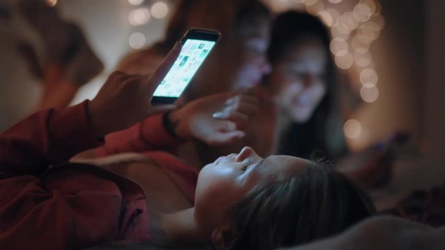 happy teenage girls lying on bed using smartphone texting on social media browsing online having fun sleepover on weekend night