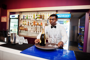 African american bartender at bar holding champagne with glasses on tray.