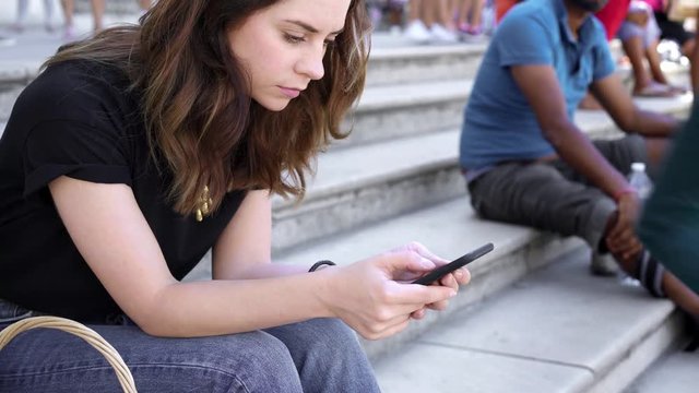 Real time shot of woman with smartphone sitting on the stairs in Paris texting. Young woman with dark hair wearing jeans and black t-shirt.