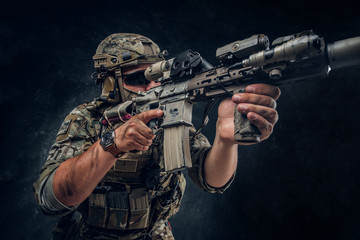 Military man in full equipment with wach on his hand is holding machine gun while posing for photographer over dark background.
