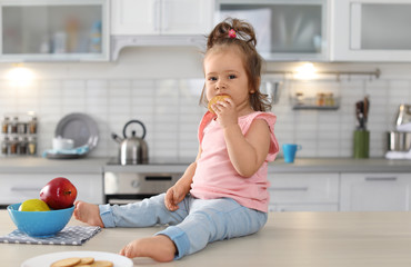 Adorable little baby girl eating tasty cookie on table in kitchen