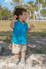 A toddler stands upon a large rock at the park and looks to his left. The summer sun dapples through the trees provides some shade for the little boy with his hand up for balance.