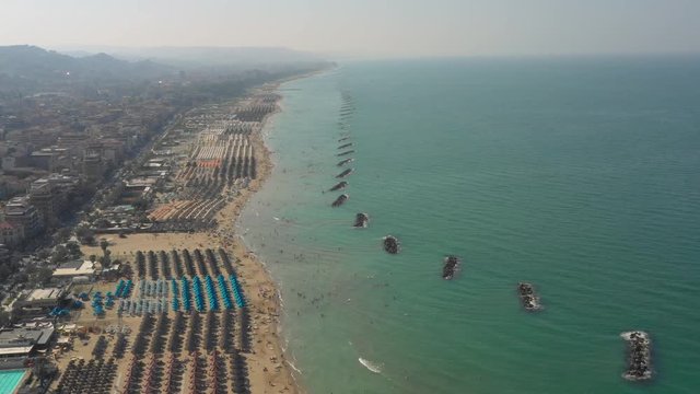 Wide drone shot of the beaches of Pescara, Italy showcasing the rock formations off the coast in the ocean, the beach and beach goer's, colorful umbrellas, buildings, cars, trees, and mountains.