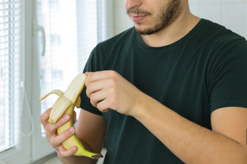 A young handsome brunette man is standing in the kitchen at home and cleans a banana.
