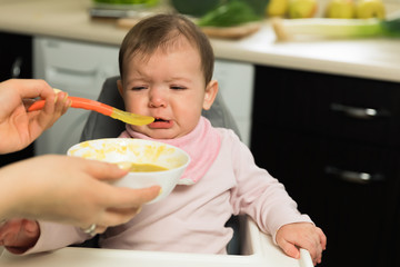 Mom feeds a baby with a spoon. The baby is crying while sitting at the baby’s feeding chair.