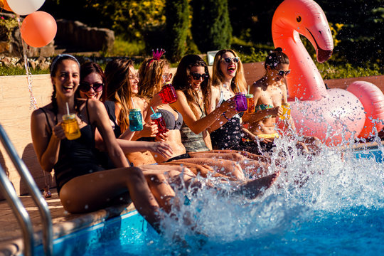Group of girlfriends at a poolside summer party sitting at the edge of a swimming pool drinking and having fun.